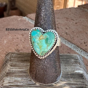 May include: A sterling silver ring featuring a heart-shaped turquoise stone. The turquoise is a vibrant teal color, set within a twisted rope-like border. The ring is displayed on a wooden stand. The text "925SilverArtsCo" is visible.