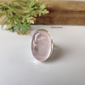 Könnte beinhalten: Ein Silberring mit einem großen, ovalen Rosenquarz-Edelstein. Der hellrosa Stein ist in einem schlichten Silberband gefasst. Der Ring wird auf einer weißen Oberfläche präsentiert, mit Grün im Hintergrund. Der Text "925SilverArtsCo" ist sichtbar.