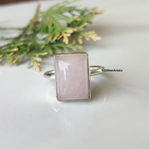May include: A silver ring featuring a rectangular, pale pink rose quartz gemstone. The ring is set against a white surface, with a sprig of green foliage in the background. The text "925SilverArtsCo" is visible.