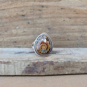 K&ouml;nnte beinhalten: Ein tropfenf&ouml;rmiger Ring mit einem polierten Ammoniten-Fossil mit braunen und beigen Spiralmustern. Der Stein ist in einen silbernen Rahmen mit einer gedrehten Kordelbord&uuml;re gefasst. Der Text 925SilverLegance ist auf der rechten Seite zu sehen.