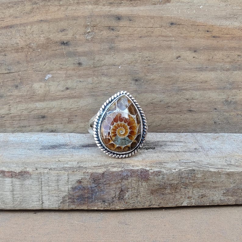 K&ouml;nnte beinhalten: Ein tropfenf&ouml;rmiger Ring mit einem polierten Ammoniten-Fossil mit braunen und beigen Spiralmustern. Der Stein ist in einen silbernen Rahmen mit einer gedrehten Kordelbord&uuml;re gefasst. Der Text 925SilverLegance ist auf der rechten Seite zu sehen.