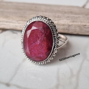 May include: A silver ring featuring a large, oval, faceted deep red gemstone set in a twisted rope-style metal border. The band is split at the sides, and the text 925SilverLegance is visible near the base of the ring setting.