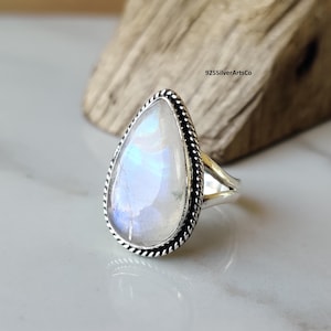 May include: A silver ring featuring a pear-shaped moonstone. The moonstone displays a milky white and blue iridescence. The ring has a twisted rope design around the stone. The ring is on a white surface with a wooden background.