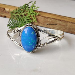 May include: A silver cuff bracelet featuring an oval, blue opal gemstone. The stone is set in a silver frame with a twisted rope design. The bracelet has decorative silver swirls on each side.