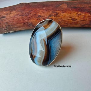 May include: A ring featuring a large, oval-shaped polished agate gemstone set in a silver-toned metal band. The stone displays concentric bands of brown, white, and translucent blue colours. The text 925SilverLegance is visible on the surface.