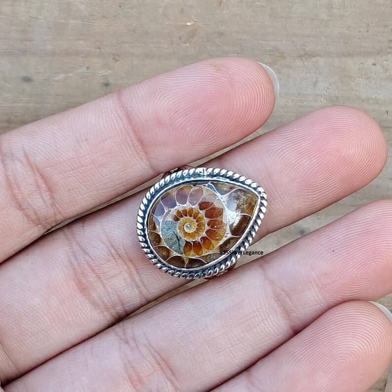 K&ouml;nnte beinhalten: Ein tropfenf&ouml;rmiger Ring mit einem polierten braun-beigen Ammonitenfossil, das in einen gedrehten silberfarbenen Metallrand gefasst ist. Das Fossil zeigt ein klares Spiralmuster mit deutlichen Kammern. Der Text 925SilverLegance ist zu sehen.