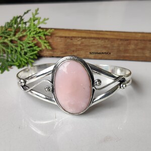 Puede incluir: Una pulsera de plata con una gema ovalada de color rosa pálido. La gema está engastada en un bisel de plata y la pulsera tiene un diseño calado decorativo. La pulsera es una joya.