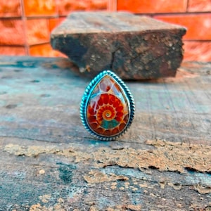 K&ouml;nnte beinhalten: Ein tropfenf&ouml;rmiger Ring mit einer polierten Ammoniten-Fossil-Einlage, die leuchtend orange, rote und t&uuml;rkisfarbene Spiralmuster aufweist. Das Fossil ist in eine silberfarbene Metallfassung mit einem gedrehten Seilrand eingefasst.
