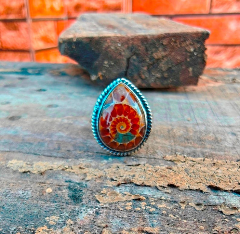 K&ouml;nnte beinhalten: Ein tropfenf&ouml;rmiger Ring mit einer polierten Ammoniten-Fossil-Einlage, die leuchtend orange, rote und t&uuml;rkisfarbene Spiralmuster aufweist. Das Fossil ist in eine silberfarbene Metallfassung mit einem gedrehten Seilrand eingefasst.