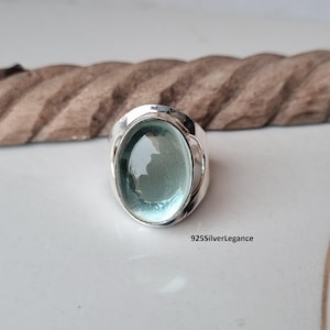 May include: A silver ring featuring a large, oval-shaped, translucent light green gemstone set in a polished metal band. The text 925SilverLegance is printed in black near the base of the ring, indicating the metal composition and brand name.