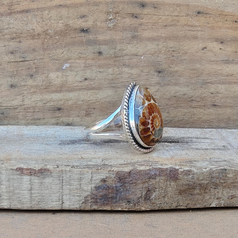 K&ouml;nnte beinhalten: Ein Silberring mit einem tropfenf&ouml;rmigen Ammoniten-Fossil-Edelstein mit Spiralmuster in Braun- und Beiget&ouml;nen. Die Fassung hat einen gedrehten Kordelrand und eine geteilte Ringschiene. Der Text 925SilverLegance ist an der Seite deutlich zu erkennen.