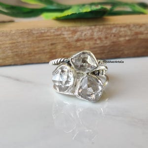Op de afbeelding: Een zilveren ring met drie heldere, facetgeslepen edelstenen. De ring heeft een gedraaide band en een clusterontwerp, met de edelstenen in een driehoekig patroon gerangschikt. De ring is geplaatst op een wit oppervlak met een houten achtergrond.