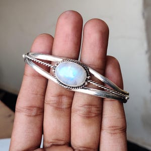 May include: A silver cuff bracelet featuring a large, oval moonstone. The moonstone has a pearly, iridescent sheen. The bracelet has a twisted rope design on the sides and around the moonstone. The bracelet is shown on a hand.