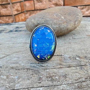 May include: Oval-shaped ring featuring a vibrant blue stone with iridescent accents. The stone is set in a silver-toned metal band with a twisted rope design around the edge. The ring is displayed on a wooden surface.