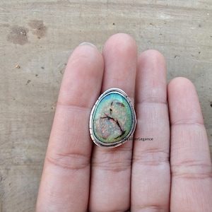 May include: A sterling silver ring featuring a large, oval-shaped opal gemstone. The opal displays a mix of colors, including green, orange, and black, with a unique crackled pattern. The ring has a detailed, textured silver band. The text "925SilverLegance" is visible.