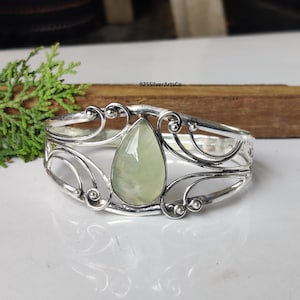 May include: A silver-toned cuff bracelet with an openwork design and a pear-shaped, pale green gemstone at the center. The bracelet has decorative swirls and the text "925SilverArtsCo".