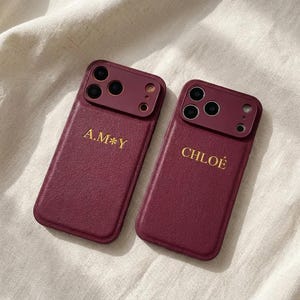 Peut inclure: Deux coques de téléphone bordeaux avec une finition texturée et un cache-objectif assorti. Chaque coque présente des lettres dorées, l'une personnalisée avec le nom A.M*Y et l'autre avec le nom CHLOÉ dans une police de caractères serif.