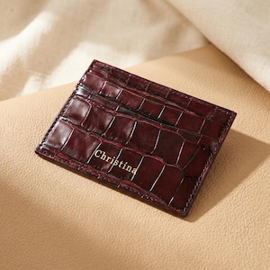 Peut inclure: Un porte-cartes rectangulaire bordeaux foncé avec une texture en cuir de crocodile brillant. La surface avant présente le nom Christina embossé en lettres dorées. L'objet comporte des coutures visibles sur les bords et plusieurs fentes.