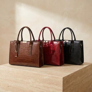 Peut inclure: Trois sacs à main structurés en marron, rouge et noir, chacun présentant une texture gaufrée façon crocodile brillante, deux poignées supérieures et une petite plaque métallique dorée rectangulaire centrée sur le bord supérieur avant.