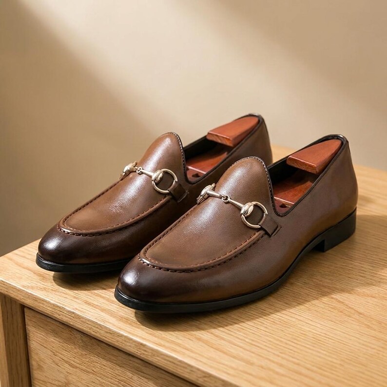 May include: A pair of brown leather loafers featuring a gold-toned metal horsebit detail across the vamp. The shoes have a smooth finish, a rounded toe, and a low black heel. Each shoe contains a wooden shoe tree insert for shape retention.