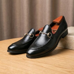 May include: A pair of black leather loafers featuring a gold-toned metal horsebit detail across the vamp. The shoes have a smooth finish, a rounded toe, and a low heel, with wooden shoe trees inserted inside to maintain their structured shape.