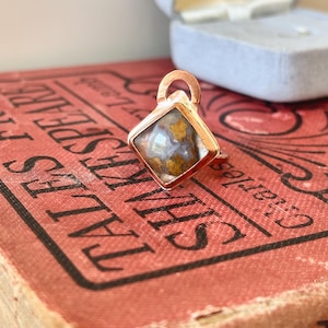 May include: A square pendant with a copper-colored frame and a stone center. The stone has a mix of blue, brown, and yellow hues. The pendant is resting on a red book with gold lettering. A blue jewelry box is in the background.