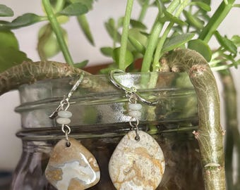 May include: A pair of handcrafted earrings featuring natural stone pendants. The earrings have a silver-colored hook and small, light-colored beads. The stone pendants are beige with brown and white patterns, showcasing a unique, organic design.