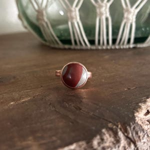 May include: A copper-colored ring with a round, reddish-brown stone and a silver-colored stripe. The ring is set on a wooden surface. A glass bottle in a macrame holder is in the background.