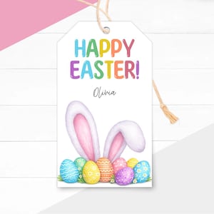 Puede incluir: Una etiqueta de regalo de Pascua blanca con las palabras "HAPPY EASTER!" en letras coloridas. Debajo del texto está el nombre "Olivia" en cursiva. La etiqueta presenta orejas de conejo y coloridos huevos de Pascua. Se adjunta un cordel natural.