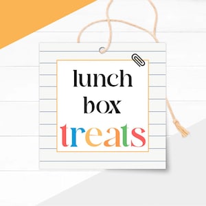 May include: Square tag with the words "lunch box treats" in black and colorful letters. The tag has a paperclip and is attached to a string. The background is white with a wood grain pattern.