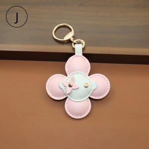 K&ouml;nnte beinhalten: Ein Schl&uuml;sselanh&auml;nger aus rosa und hellblauem Leder in einem vierbl&auml;ttrigen Blumenmuster. Die Mitte der Blume ist hellblau mit einer rosa Blume und goldenen Akzenten. Der Schl&uuml;sselanh&auml;nger hat einen goldfarbenen Ring.