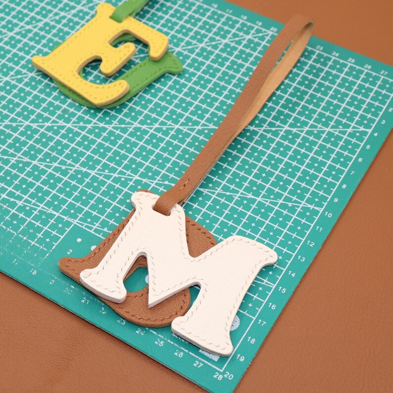 May include: A leather luggage tag with the letter "M" in white, stitched onto a brown base, and a tan strap. A green and yellow letter "E" is in the background. The tag is on a green cutting mat.
