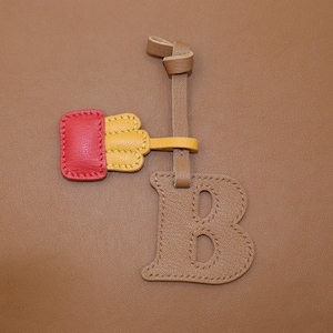 May include: A leather luggage tag with a tan background. The tag features a red and yellow accent piece, a tan strap, and a tan letter "B" with stitched details. The tag is likely for a suitcase or bag.