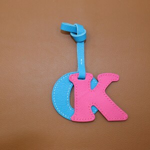 May include: A leather keychain featuring the letters "OK". The "O" is light blue, and the "K" is pink, with white stitching. The keychain has a blue strap with a knotted top.
