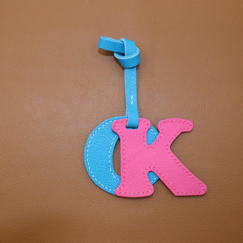 May include: A leather keychain featuring the letters "OK". The "O" is light blue, and the "K" is pink, with white stitching. The keychain has a blue strap with a knotted top.