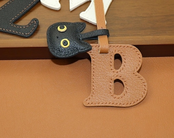 May include: Leather letter "B" in a light brown color with a cat-shaped charm in black. The letter has a stitched border and two cut-out ovals. The cat charm has yellow eyes. Other letters are visible in the background.