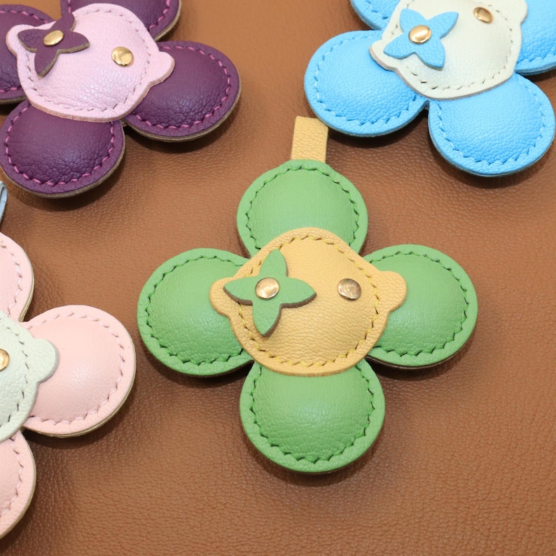 K&ouml;nnte beinhalten: Nahaufnahme von mehreren ledernen bl&uuml;tenf&ouml;rmigen Accessoires in verschiedenen Farben. Die Blumen haben vier Bl&uuml;tenbl&auml;tter und ein zentrales Design mit einer kleinen Schlaufe oben. Zu den Farben geh&ouml;ren Rosa, Lila, Gr&uuml;n, Gelb und Blau mit goldfarbenen Akzenten.