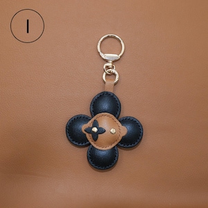 K&ouml;nnte beinhalten: Ein Leder-Schl&uuml;sselanh&auml;nger mit einem vierbl&auml;ttrigen Blumenmuster. Die Bl&uuml;tenbl&auml;tter sind schwarz, und die Mitte ist hellbraun mit einem goldfarbenen Akzent. Der Schl&uuml;sselanh&auml;nger hat einen goldfarbenen Ring und Verschluss.