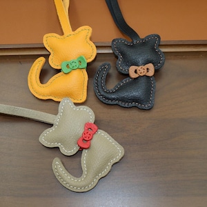May include: Three cat-shaped leather charms with straps. One is yellow with a green bow, one is black with a tan bow, and one is beige with a red bow. Each charm features visible stitching along the edges and a small bow at the neck area.