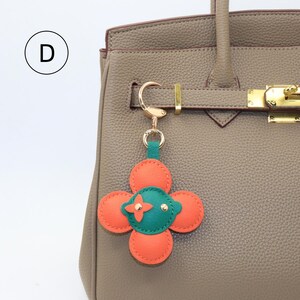 K&ouml;nnte beinhalten: Ein bl&uuml;tenf&ouml;rmiger Schl&uuml;sselanh&auml;nger mit orangefarbenen und gr&uuml;nen Akzenten h&auml;ngt an einer hellbraunen Handtasche. Der Schl&uuml;sselanh&auml;nger hat vier orangefarbene Bl&uuml;tenbl&auml;tter, die einen gr&uuml;nen Mittelpunkt mit einem roten Kreuz umgeben. Die Handtasche hat goldfarbene Hardware.