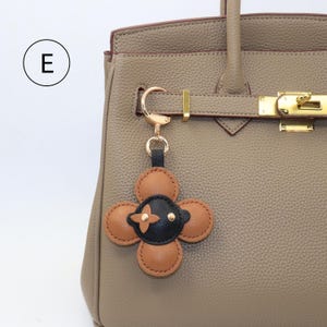 K&ouml;nnte beinhalten: Ein lederner, blumenf&ouml;rmiger Schl&uuml;sselanh&auml;nger in Braun und Schwarz mit goldfarbener Hardware, befestigt an einer taupefarbenen Handtasche. Der Schl&uuml;sselanh&auml;nger hat eine schwarze Mitte mit einem goldenen Kreuzdesign und ist ein dekoratives Accessoire.