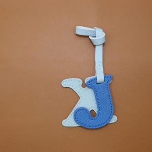 May include: A light blue and blue leather letter charm with a knotted strap. The charm features the letters "J" and "K" stitched together. The background is a solid brown color.