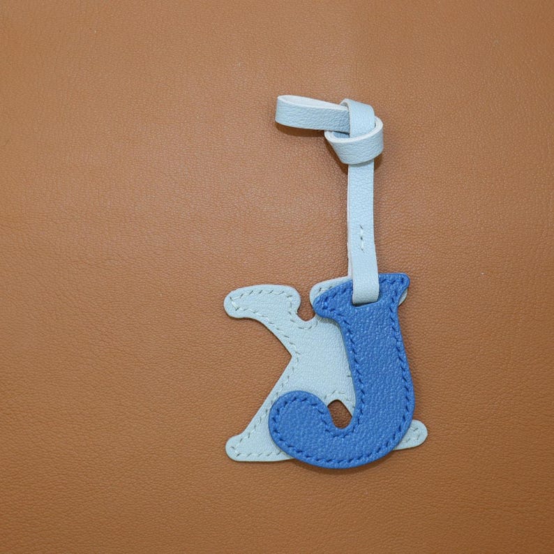 May include: A light blue and blue leather letter charm with a knotted strap. The charm features the letters "J" and "K" stitched together. The background is a solid brown color.