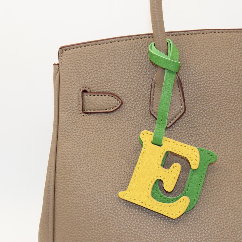 May include: A close-up of a taupe leather handbag with a decorative leather charm. The charm is in the shape of the letter "E", with yellow and green color blocking. The handbag has a top handle and a metal clasp.