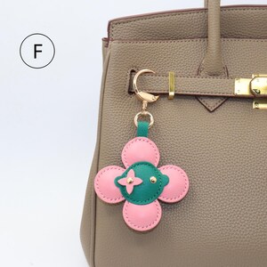 K&ouml;nnte beinhalten: Ein rosa-gr&uuml;ner, bl&uuml;tenf&ouml;rmiger Schl&uuml;sselanh&auml;nger mit goldfarbener Hardware. Der Anh&auml;nger ist an einer taupefarbenen Lederhandtasche befestigt. Die Blume hat vier rosa Bl&uuml;tenbl&auml;tter und eine gr&uuml;ne Mitte mit goldenen Akzenten. Die Handtasche hat einen goldenen Verschluss.