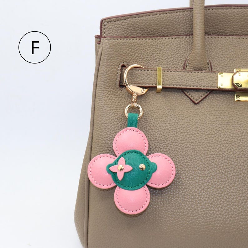 K&ouml;nnte beinhalten: Ein rosa-gr&uuml;ner, bl&uuml;tenf&ouml;rmiger Schl&uuml;sselanh&auml;nger mit goldfarbener Hardware. Der Anh&auml;nger ist an einer taupefarbenen Lederhandtasche befestigt. Die Blume hat vier rosa Bl&uuml;tenbl&auml;tter und eine gr&uuml;ne Mitte mit goldenen Akzenten. Die Handtasche hat einen goldenen Verschluss.