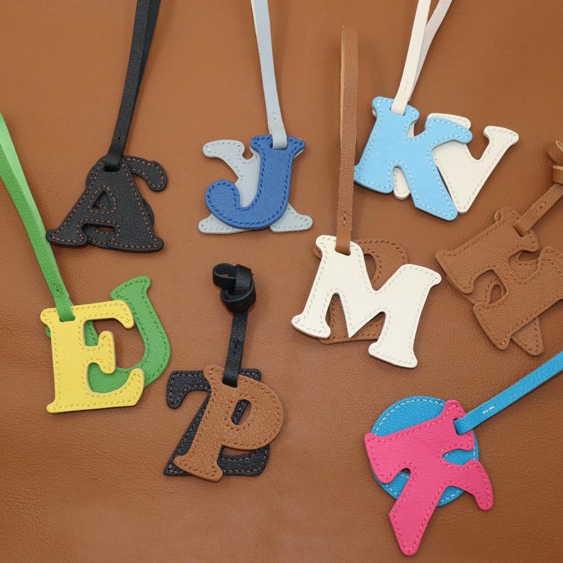 May include: A collection of leather letter charms in various colors, including black, blue, white, brown, green, yellow, and pink. Each letter has a matching strap for attaching to a bag or keychain. The letters are stitched with contrasting thread.