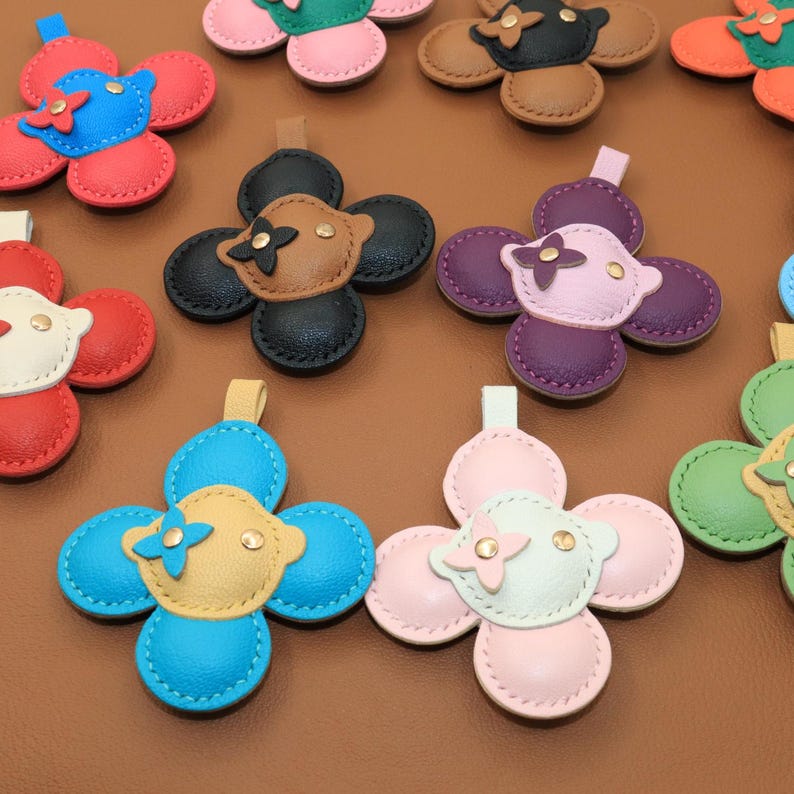 K&ouml;nnte beinhalten: Lederanh&auml;nger in Blumenform in verschiedenen Farben wie Rot, Blau, Rosa und Gr&uuml;n. Jeder Anh&auml;nger hat eine kontrastierende Mitte mit einem kleinen dekorativen Element und einer Schlaufe zur Befestigung.