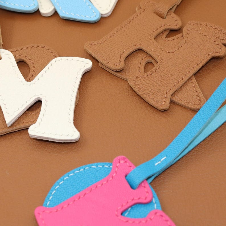 May include: Close-up of leather letter charms in white, tan, and blue. The letter "M" is white, and the letter "H" is tan. A pink and blue charm is in the foreground. The charms are stitched with white thread.