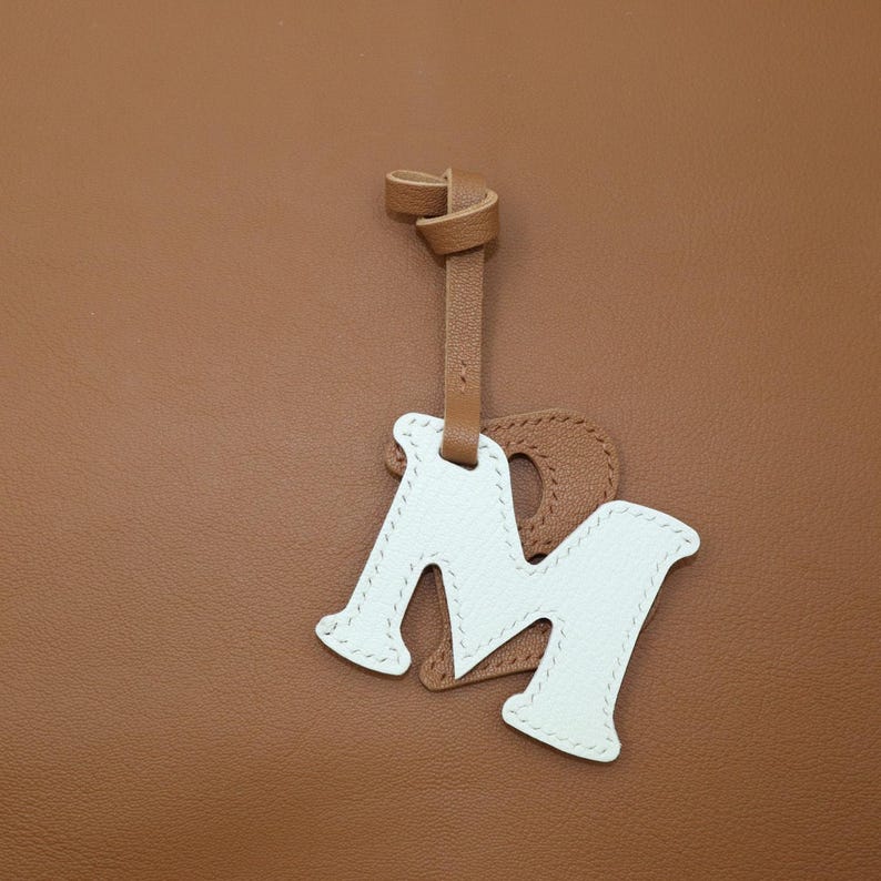 May include: A leather bag charm featuring a white letter "M" with a brown leather backing and a matching strap. The charm is stitched with white thread and hangs against a brown leather background.